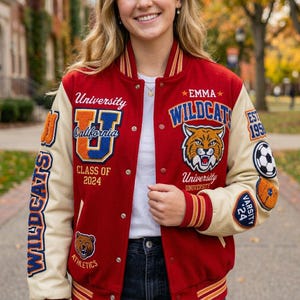Op de afbeelding: Een rode en crèmekleurige varsityjas met de tekst "WILDCATS" en "University California" op de voorkant. De jas heeft een tijgermascotte-patch en de tekst "CLASS OF 2024". De mouwen zijn crèmekleurig.