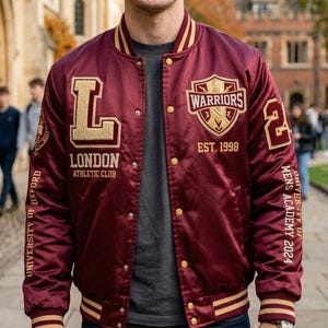 Könnte beinhalten: Burgunderfarbene Satin-Bomberjacke mit goldenen Akzenten. Mit den Schriftzügen "LONDON ATHLETIC CLUB" und "WARRIORS EST. 1998". Die Jacke hat einen gerippten Kragen, Bündchen und Saum mit goldenen Streifen. Die Ärmel tragen die Aufschrift "UNIVERSITY OF OXFORD" und "MENS ACADEMY 2024".
