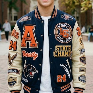 Pode incluir: Casaco universitário azul marinho e creme com detalhes em laranja e branco. Apresenta patches com textos como "REBELS", "Legacy Athletics" e "State Champ". Inclui gráficos de águia e pantera.