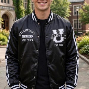 May include: A black varsity jacket with white striped trim on the collar, cuffs, and hem. The front features white embroidery reading City University Athletics and a large U logo with the text Lions Est. 2023. It has a snap-button closure.