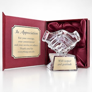 Könnte beinhalten: Eine klare Glasskulptur in Form eines Händedrucks auf einem facettierten Kristallsockel mit einer goldenen Plakette mit der Aufschrift With respect and gratitude. Sie wird in einer offenen burgunderroten Schachtel mit einer goldenen Plakette präsentiert, auf der steht: In Appreciation For your courage, your commitment and your service to others. Thank you for everything you do.