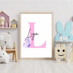 Könnte beinhalten: Ein rosa Buchstabe L mit dem Namen "Liya" in Kursivschrift. Ein lilafarbener Schmetterling mit rosa und weißen Flügeln befindet sich auf der linken Seite des Buchstabens. Der Druck ist in einem hellen Holzrahmen gerahmt.