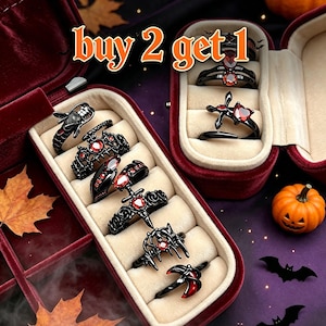 May include: A collection of black metal rings featuring red gemstone accents, displayed in a velvet jewelry box. The rings include designs with hearts, crosses, and intricate patterns. The text "buy 2 get 1" is visible at the top of the image.