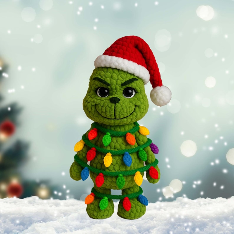 May include: A green crocheted character wearing a red Santa hat with a white trim and a string of colorful Christmas lights. The character has a smiling face and is standing in the snow, perfect for Christmas.