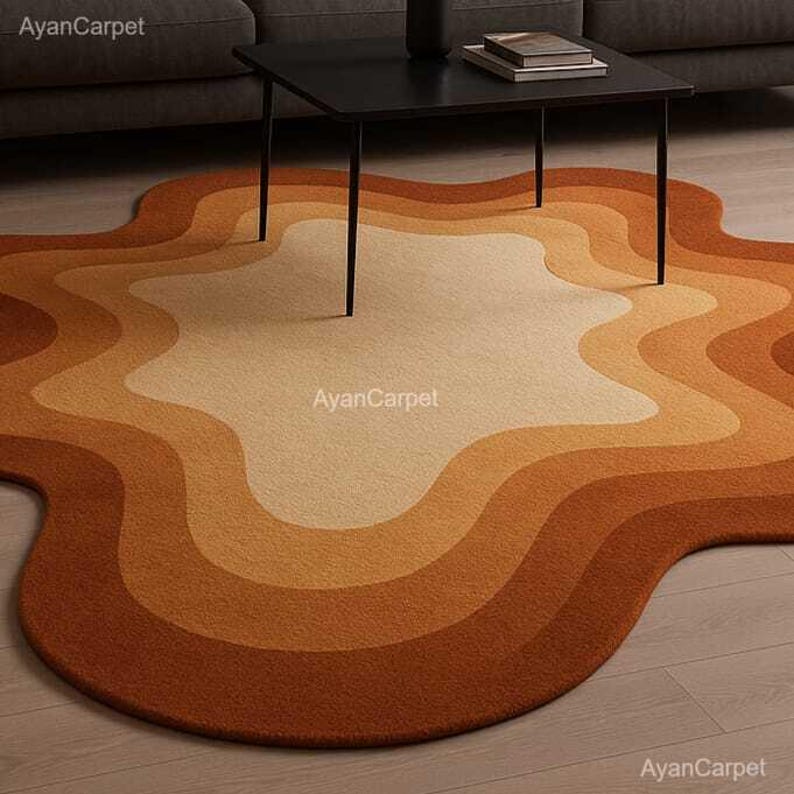 May include: A large, abstract area rug with a wavy, organic shape. The rug has a light beige center with concentric rings of brown, orange, and rust. The rug is shown on a light wood floor with a black coffee table in the background.