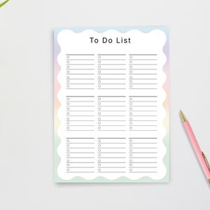 May include: A pastel-colored To Do List notepad with a wavy border. The notepad has a light blue, pink, and green gradient. The words "To Do List" are printed at the top. A pink pen with gold accents rests on the white surface.