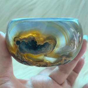 May include: A polished, translucent bangle bracelet crafted from banded agate stone. The circular accessory features swirling patterns of amber, golden yellow, and deep blue-grey tones, with a smooth, glossy finish that highlights the natural mineral layers.
