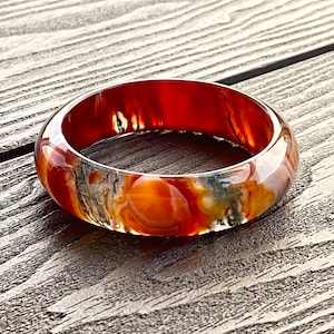 Könnte beinhalten: Ein durchscheinendes, rundes Armreif-Armband mit marmoriertem Aussehen. Das Armband weist Farbtöne von Rot, Orange, Gelb und Grau auf. Der Schmuck liegt auf einer Holzoberfläche.