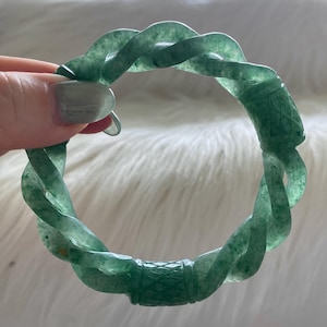 Op de afbeelding: Een doorschijnende groene armband met een gedraaid, touwachtig ontwerp. Het cirkelvormige accessoire heeft een gladde, gepolijste afwerking met een duidelijk ruitpatroon dat in een deel van de band is gegraveerd voor textuur.