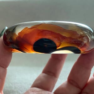 Könnte beinhalten: Ein glatter, abgerundeter Armreif aus klarem Harz oder Glas mit internen, wirbelnden Mustern in tiefen Bernstein-, gebrannten Orange- und Dunkelbrauntönen, die einen geschichteten, organischen Effekt innerhalb des transparenten Materials erzeugen.