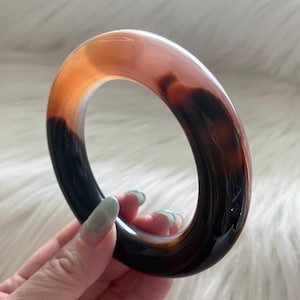 May include: A smooth, polished bangle bracelet crafted from banded agate stone. The circular jewelry piece features a gradient of translucent orange and deep, opaque black tones with natural swirling patterns throughout the polished surface.