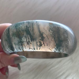 May include: A smooth, polished bangle bracelet crafted from translucent moss agate stone. The material features a milky, semi-clear base with intricate, dark green, tree-like dendritic inclusions that resemble a forest landscape.