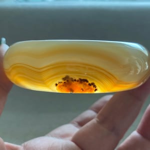 May include: A smooth, polished bangle bracelet crafted from translucent yellow and cream-colored agate. The stone features distinct concentric banding patterns and a prominent, dark, textured inclusion near the bottom edge of the circular band.