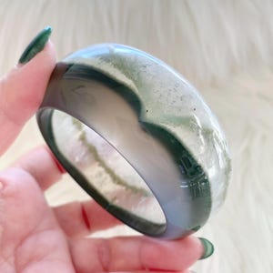 May include: A smooth, polished bangle bracelet crafted from translucent stone featuring distinct bands of deep forest green and cloudy white. The circular accessory has a glossy, reflective surface that highlights the natural mineral inclusions and colour variations within the stone material.