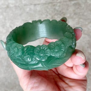 Puede incluir: Un brazalete rígido de color verde translúcido con un motivo floral tallado que recorre toda la superficie exterior. El accesorio circular tiene una banda interior lisa y un patrón en relieve detallado de flores y hojas en tono verde.