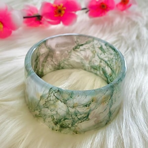 Op de afbeelding: Een doorschijnende, brede armband met groene, mosachtige insluitsels. De armband ligt tegen een witte, donzige achtergrond, met roze bloemen op de achtergrond. Het ontwerp van de armband is organisch en natuurlijk.
