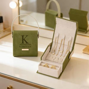 以下が含まれることがあります： 白い内装のオリーブグリーンのジュエリーボックス。金色のネックレスと指輪が入っています。ボックスにはハンドルがあり、前面には「Kirsten」という名前が印刷されています。