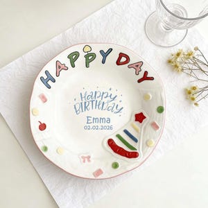 以下が含まれることがあります： ピンクの縁取りのある白いセラミックプレート。カラフルな文字とイラストで装飾されています。プレートには縁に「HAPPY DAY」、中央に「Happy Birthday Emma 02.02.2026」と書かれており、パーティーハットのイラストが描かれています。