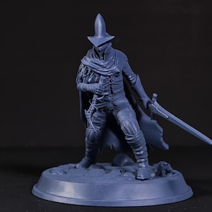 Könnte beinhalten: Blaue Figur einer Figur in einem langen Mantel, Hut und mit einem Schwert. Die Figur steht auf einem kreisförmigen Sockel. Die Figur hat eine Maske und trägt einen Umhang. Die Figur befindet sich in einer Kampfhaltung.