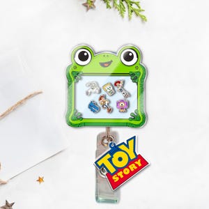 May include: A green frog-shaped badge reel featuring a clear window with Toy Story character charms. The reel has a clear plastic clip and a "Toy Story" logo charm. The background is white with a sprig of greenery.