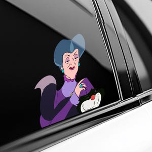 May include: A vinyl decal sticker featuring a cartoon illustration of a woman with blue hair wearing a purple dress and teal jewelry, holding a black and white cat with a red nose. The sticker is applied to the dark tinted window of a vehicle.