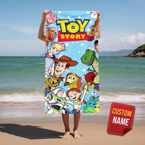 May include: A light blue beach towel featuring characters from the animated film "Toy Story." The towel displays Woody, Buzz Lightyear, and other characters against a blue sky background. A red tag with the words "CUSTOM NAME" is in the bottom right corner.