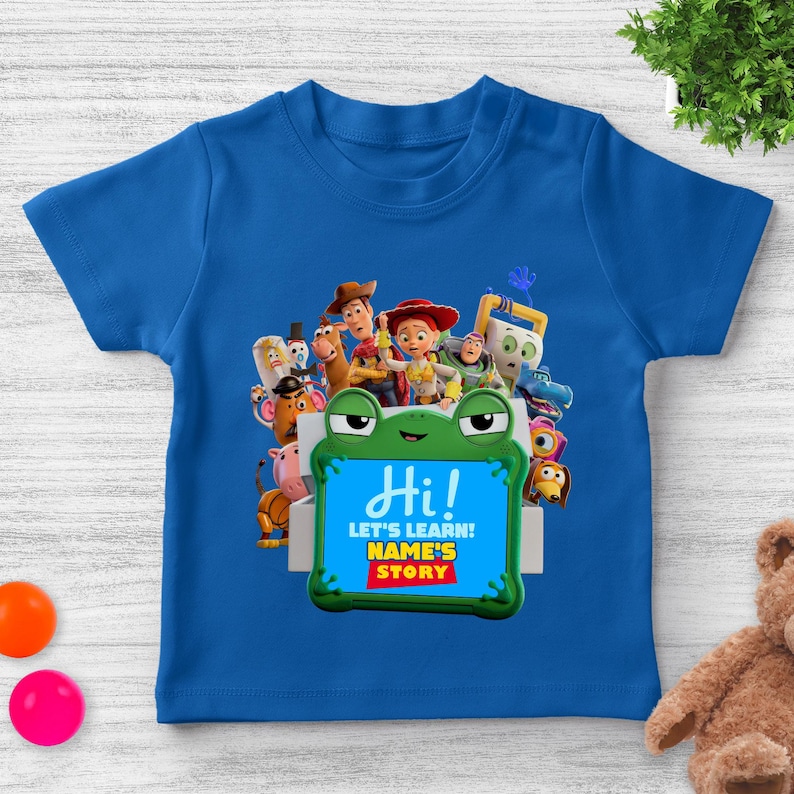 May include: A blue t-shirt with characters from "Toy Story." The shirt has a green frog-shaped sign with the words "Hi!" and "LET'S LEARN! NAME'S STORY" in white and red text. The shirt is designed for children.
