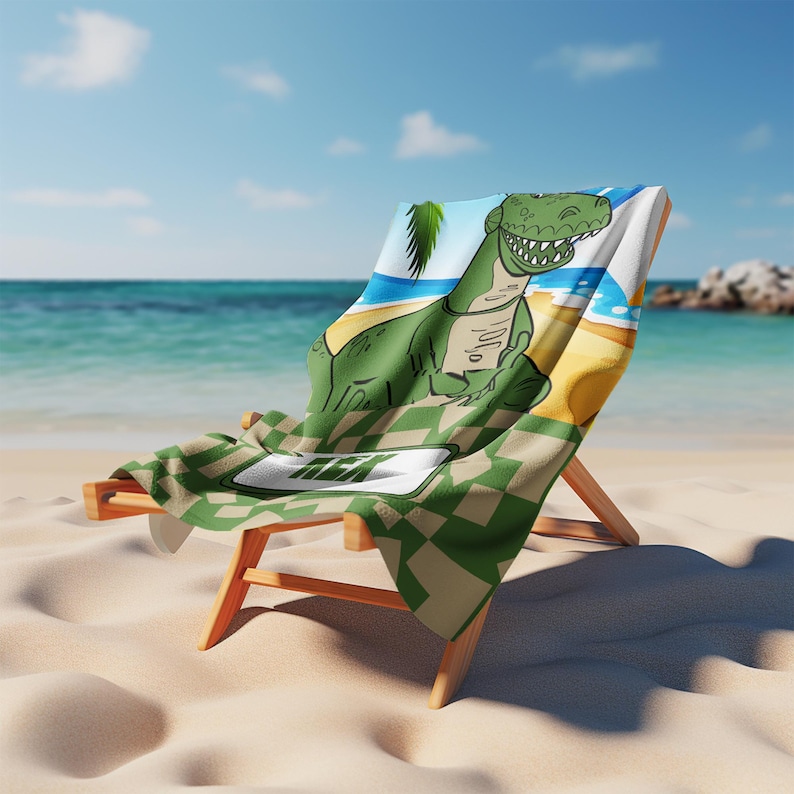 May include: A beach towel draped over a wooden lounge chair featuring a green cartoon dinosaur illustration on a beach background with a checkered pattern at the bottom and the text REX printed in a white rectangular frame on the lower section.