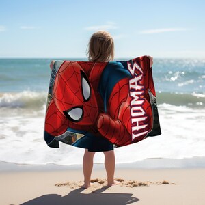 May include: A red and blue Spider-Man beach towel with the name "THOMAS" printed vertically in white. The towel is held up by a person on a sandy beach with the ocean in the background.