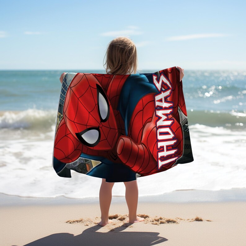 May include: A red and blue Spider-Man beach towel with the name "THOMAS" printed vertically in white. The towel is held up by a person on a sandy beach with the ocean in the background.