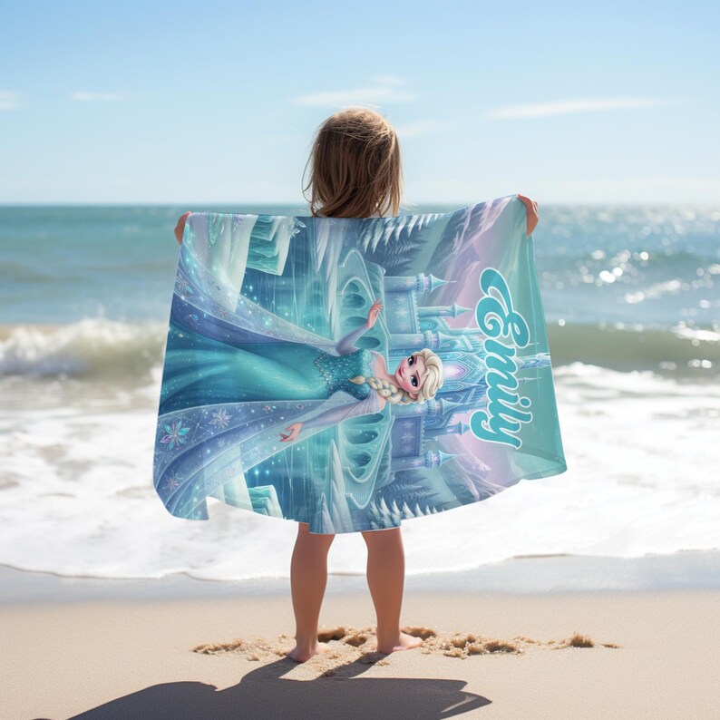 May include: A rectangular beach towel featuring a printed illustration of a blonde character in a blue gown standing in front of an ice castle. The name Emily is printed in a light blue script font on the right side of the towel, which has blue, white, and purple tones.