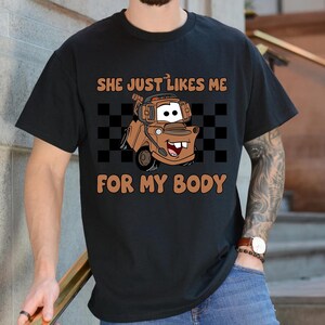 Puede incluir: Una camiseta negra de manga corta con un gr&aacute;fico de una gr&uacute;a marr&oacute;n con auriculares. Detr&aacute;s del veh&iacute;culo hay un patr&oacute;n de cuadros negros y blancos. El texto "SHE JUST LIKES ME FOR MY BODY" est&aacute; impreso en color naranja arriba y abajo.