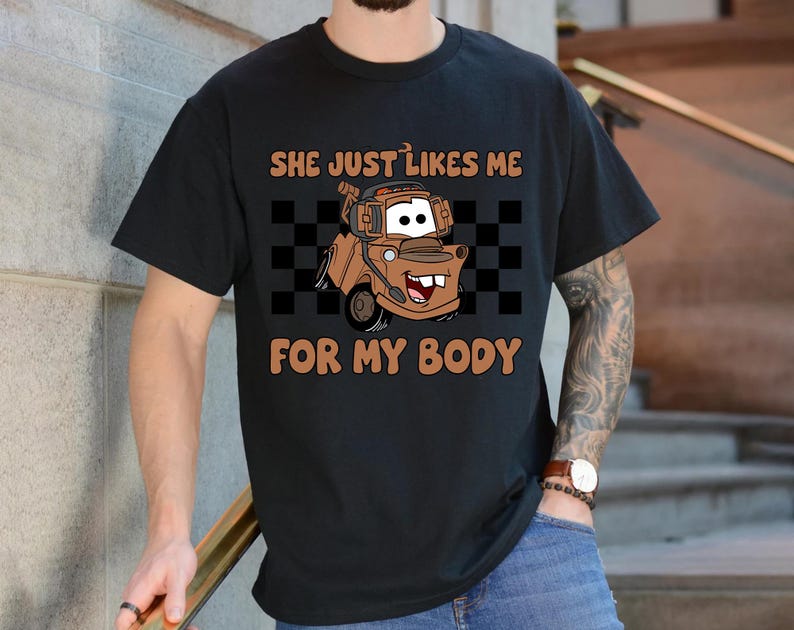 Puede incluir: Una camiseta negra de manga corta con un gr&aacute;fico de una gr&uacute;a marr&oacute;n con auriculares. Detr&aacute;s del veh&iacute;culo hay un patr&oacute;n de cuadros negros y blancos. El texto "SHE JUST LIKES ME FOR MY BODY" est&aacute; impreso en color naranja arriba y abajo.