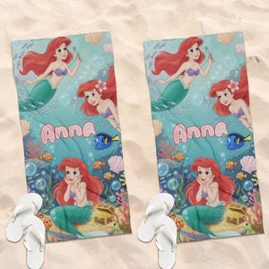 Peut inclure: Deux serviettes de plage avec un motif de sir&egrave;ne et le nom "Anna" en rose. Les serviettes ont un fond turquoise avec des illustrations de poissons, de coquillages et de coraux. Des tongs blanches sont au premier plan.