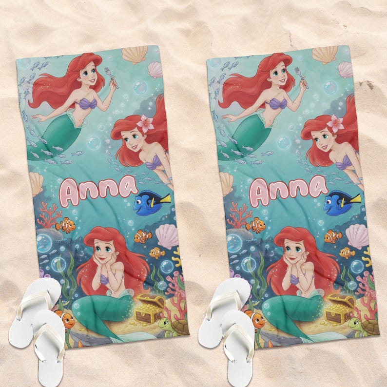 Peut inclure: Deux serviettes de plage avec un motif de sir&egrave;ne et le nom "Anna" en rose. Les serviettes ont un fond turquoise avec des illustrations de poissons, de coquillages et de coraux. Des tongs blanches sont au premier plan.
