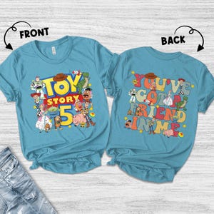 K&ouml;nnte beinhalten: Zwei hellblaue T-Shirts mit Toy Story-Designs. Das vordere Shirt zeigt "Toy Story 5" mit Charakteren. Das hintere Shirt hat "You've Got a Friend in Me" mit Charakteren. Ein farbenfrohes Design f&uuml;r Fans des Animationsfilms.