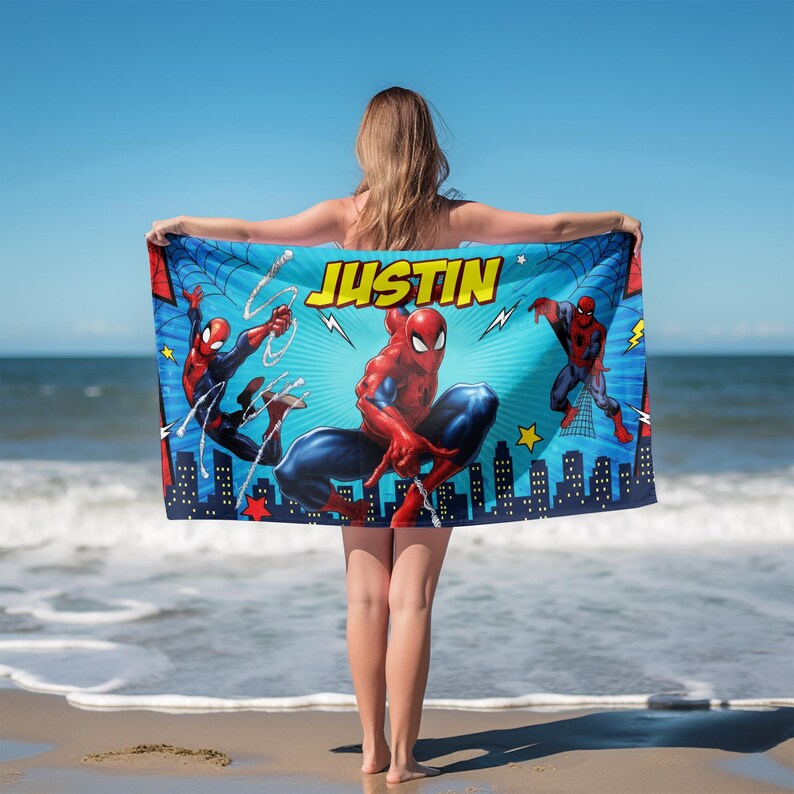 Op de afbeelding: Een blauwe strandlaken met de superheld Spider-Man in actieposes. De handdoek heeft de naam "JUSTIN" in gele blokletters. De achtergrond bevat een stadsgezicht en een webdesign.