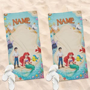 May include: Two beach towels featuring characters from The Little Mermaid, including Ariel. The towels have a light blue border with the word "NAME" in gold. White flip-flops are in the foreground, on a sandy surface.