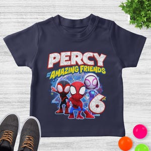 May include: A navy blue short-sleeve t-shirt featuring a graphic print of three cartoon superheroes. The text reads PERCY and his AMAZING FRIENDS above the characters, with a large number 6 positioned to the right of the figures.
