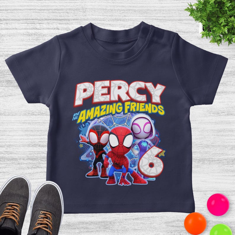 May include: A navy blue short-sleeve t-shirt featuring a graphic print of three cartoon superheroes. The text reads PERCY and his AMAZING FRIENDS above the characters, with a large number 6 positioned to the right of the figures.