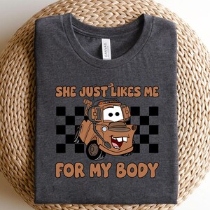 Puede incluir: Una camiseta gris jaspeado oscuro con un gr&aacute;fico de una gr&uacute;a de dibujos animados de color marr&oacute;n. El texto encima y debajo del cami&oacute;n dice: "SHE JUST LIKES ME FOR MY BODY". Un patr&oacute;n de cuadros blanco y negro se sit&uacute;a tras la ilustraci&oacute;n.