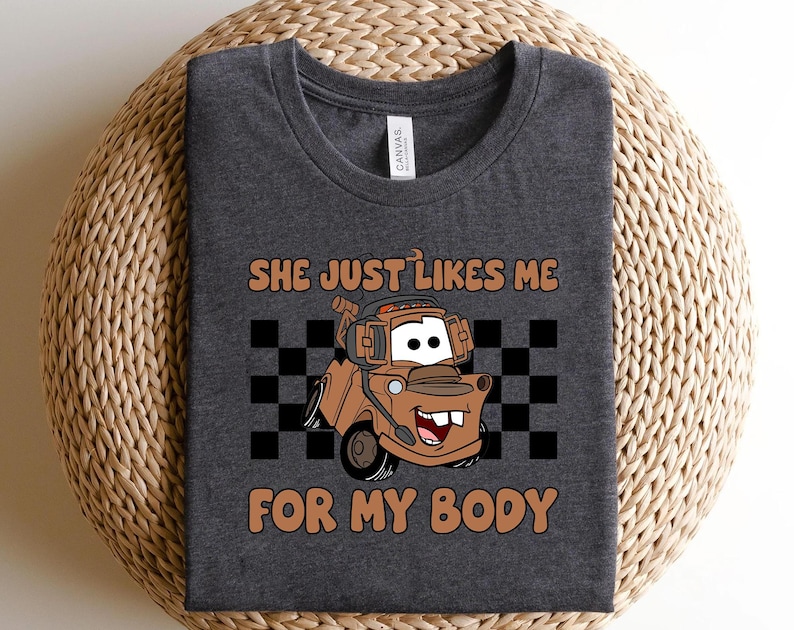 Puede incluir: Una camiseta gris jaspeado oscuro con un gr&aacute;fico de una gr&uacute;a de dibujos animados de color marr&oacute;n. El texto encima y debajo del cami&oacute;n dice: "SHE JUST LIKES ME FOR MY BODY". Un patr&oacute;n de cuadros blanco y negro se sit&uacute;a tras la ilustraci&oacute;n.
