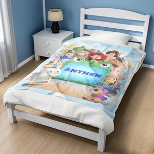 May include: A light blue blanket featuring a colorful graphic print of various animated characters surrounding a central green frog-shaped frame with the word ANTHEN printed in bold blue capital letters across the center of the design.