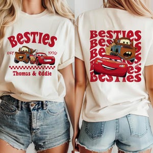 Może przedstawiać: Kremowa koszulka z napisem "Besties" w czerwonym skrypcie i grafiką samochod&oacute;w z kresk&oacute;wek. Prz&oacute;d koszulki zawiera napis "Thomas & Eddie" i "1996". Tył koszulki r&oacute;wnież zawiera napis "Besties" i grafikę samochod&oacute;w.
