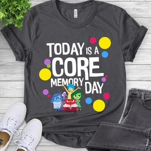 May include: Dark grey t-shirt with the text "TODAY is a CORE MEMORY DAY" in white. The shirt features cartoon characters and colourful polka dots. The shirt is paired with white trainers and dark grey jeans.