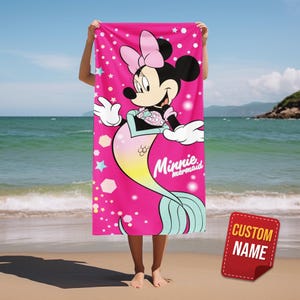 Pode incluir: Uma toalha de praia rosa com Minnie Mouse como sereia. A toalha tem um grande gr&aacute;fico da Minnie com um la&ccedil;o rosa, uma cauda de sereia e as palavras "Minnie mermaid". O fundo inclui estrelas.