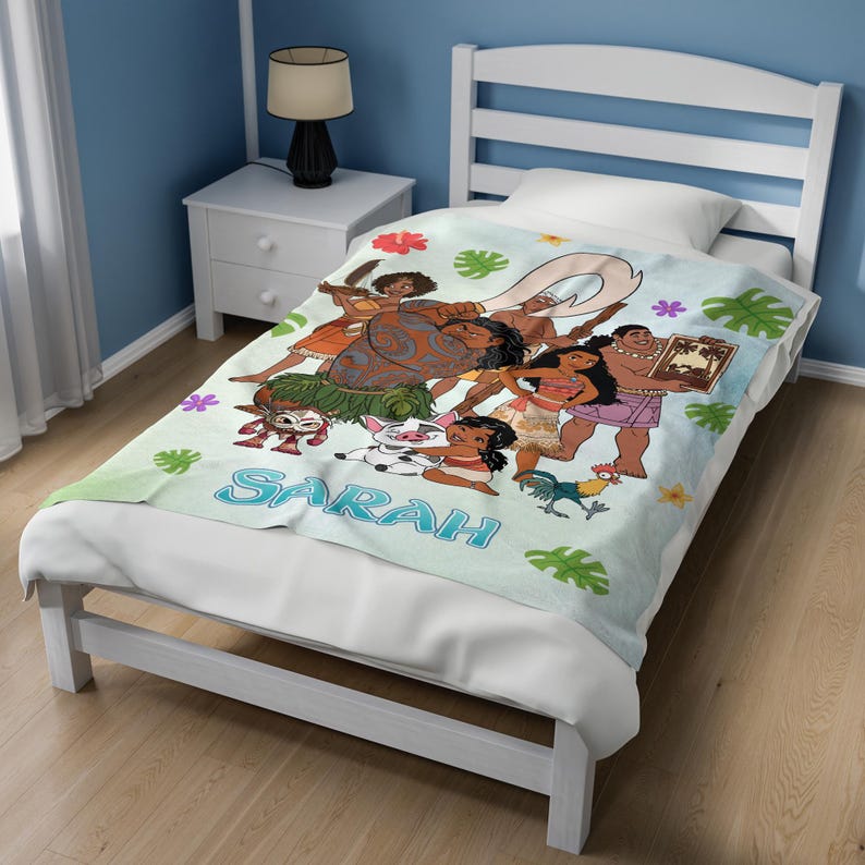 May include: A light blue fleece blanket featuring a colorful Moana-themed graphic print with characters including Moana, Maui, Pua, and Heihei, accented by tropical flowers and green leaves, with the name Sarah printed in large blue letters.
