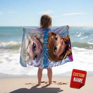 Pode incluir: Uma toalha de praia com Elsa e Anna de Frozen, com o nome "Sophia" impresso verticalmente em azul. A toalha &eacute; segurada por uma pessoa em uma praia de areia com o oceano ao fundo. Um adesivo vermelho "Custom Name" est&aacute; no canto inferior direito.