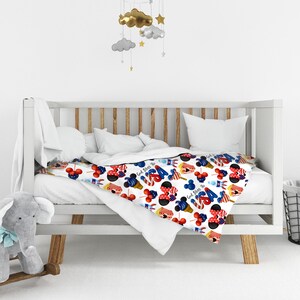 Puede incluir: Una manta de beb&eacute; blanca con un estampado colorido de cabezas de Mickey Mouse, conos de helado, cohetes y el n&uacute;mero 4 en rojo, blanco y azul. La manta est&aacute; sobre una cuna blanca.