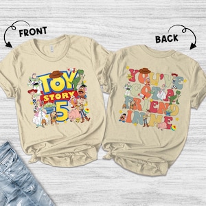 K&ouml;nnte beinhalten: Zwei beigefarbene T-Shirts mit den Aufdrucken "TOY STORY 5" und "YOU'VE GOT A FRIEND IN ME". Das vordere Shirt zeigt verschiedene Toy Story-Charaktere. Das hintere Shirt hat die gleichen Charaktere und Text in bunten Buchstaben.